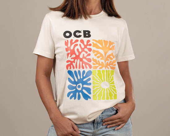A person wearing a white OCB branded t-shirt with a graphic design consisting of colorful abstract shapes in orange, blue, and yellow on the front.